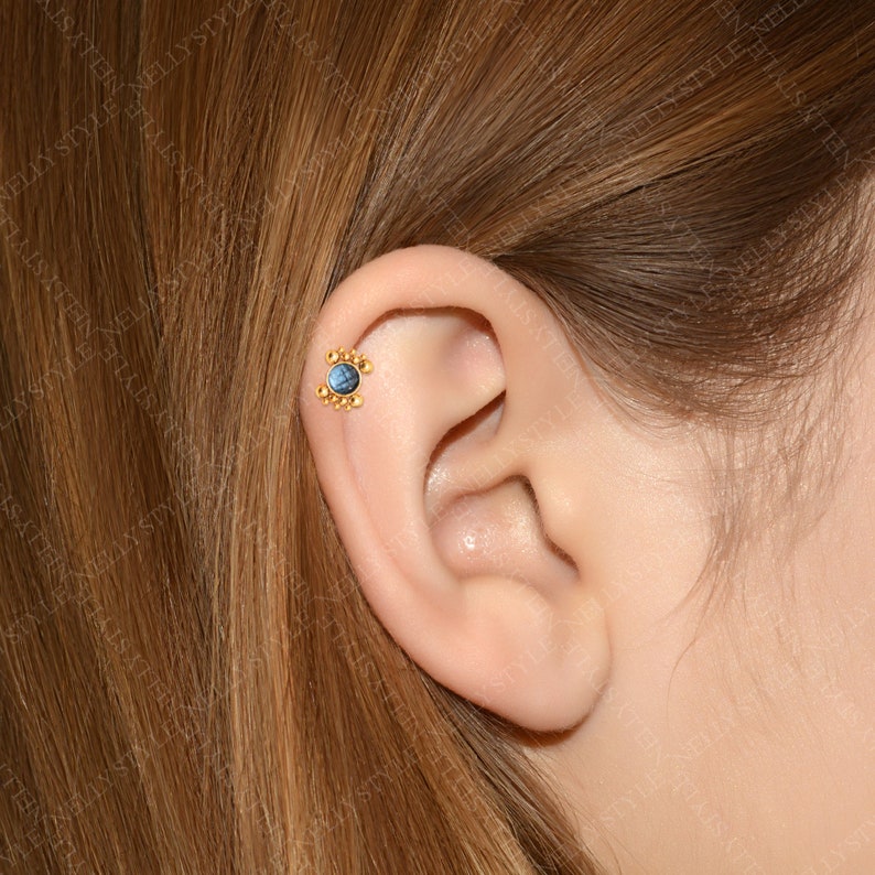 May include: A gold ear piercing with a blue gemstone in the center and a halo of small gold beads around it. The piercing is in a person's ear.