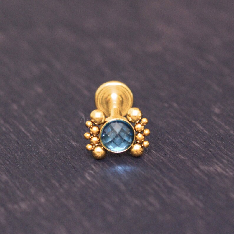 May include: Gold body jewelry with a blue gemstone and a cluster of small gold beads around the stone.