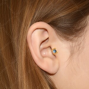 May include: A close-up of a person's ear with a gold and blue gemstone stud earring. The earring has a small, round blue gemstone surrounded by a halo of gold beads.