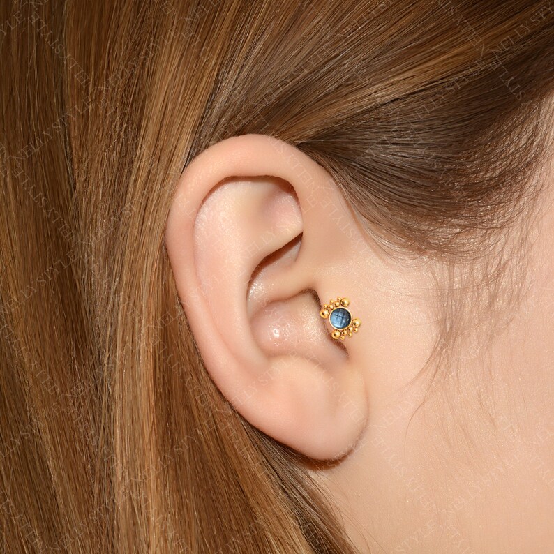 May include: A close-up of a person's ear with a gold and blue gemstone stud earring. The earring has a small, round blue gemstone surrounded by a halo of gold beads.