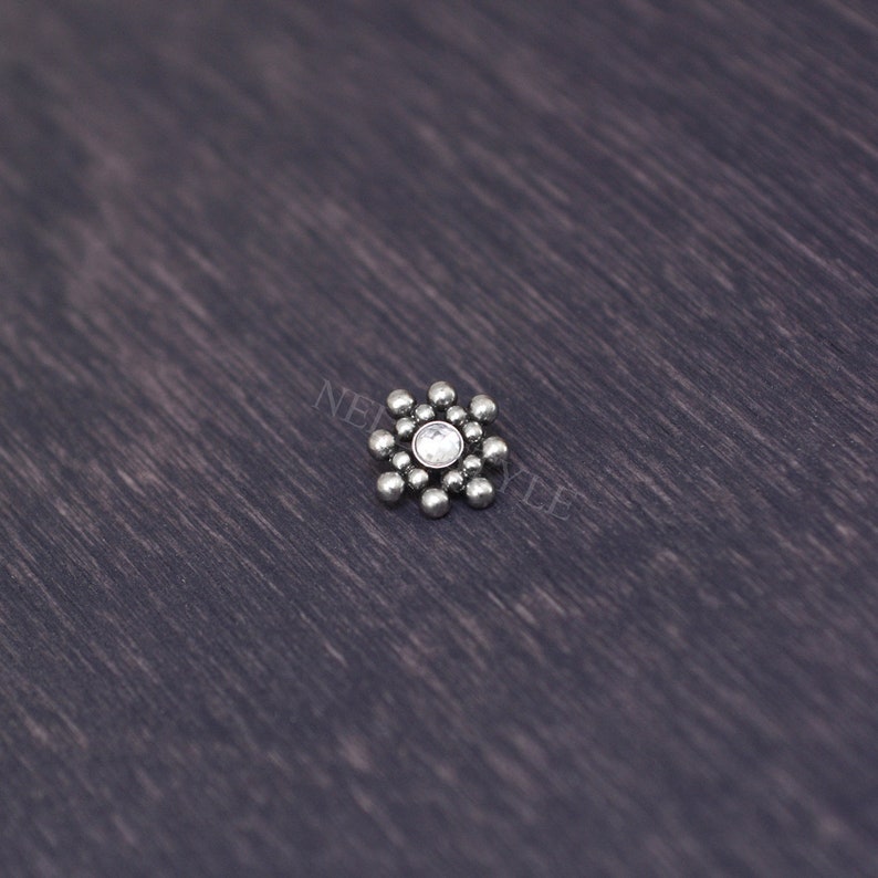 May include: A silver body jewelry piece with a clear crystal in the center and a cluster of small silver beads around it.