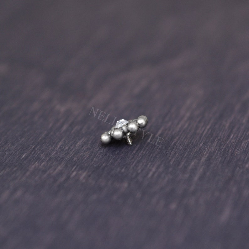 May include: A silver body jewelry piece with a clear gem in the center and five silver balls surrounding it. The jewelry piece has a screw-on back.