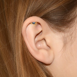 May include: A close-up of a person's ear with a gold ear piercing featuring a small blue gemstone and a row of gold beads.