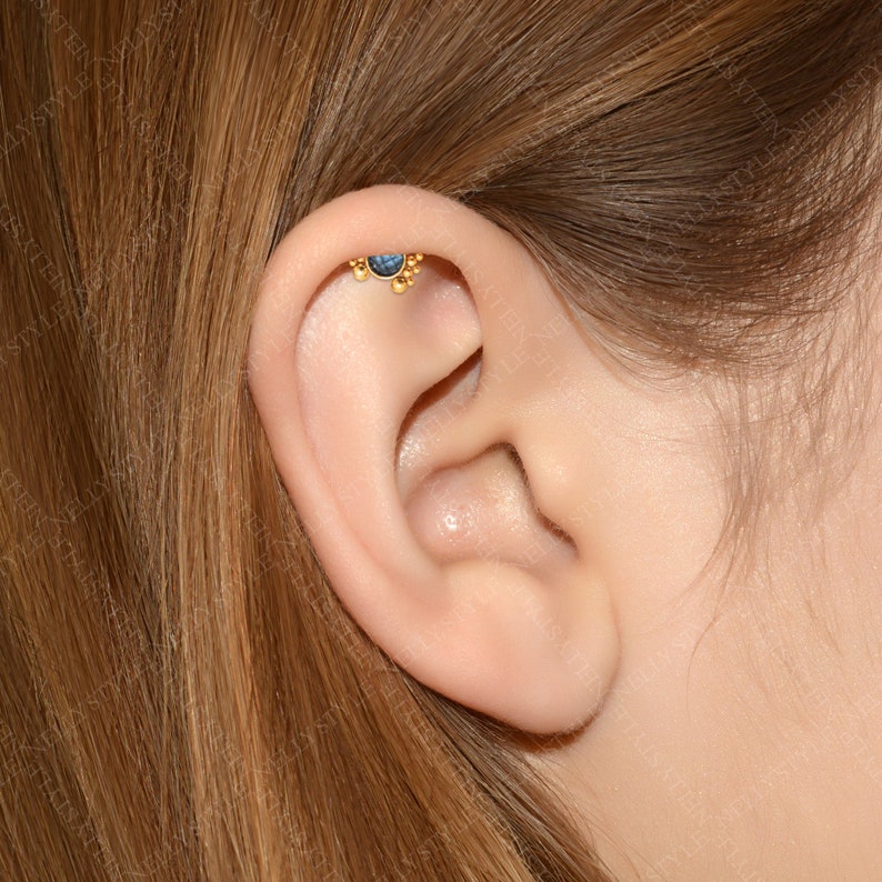 May include: A close-up of a person's ear with a gold ear piercing featuring a small blue gemstone and a row of gold beads.