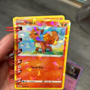 May include: A Vulpix Pokémon trading card, featuring a fox-like creature with orange fur and a fiery tail, is held in a red spiral-bound notebook. The card has a holographic design and the text "Smash Kick" with a value of 10.