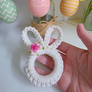 May include: A handmade white macrame bunny decoration, featuring a small pink rose. The bunny is held in a hand, with pastel-colored Easter eggs hanging in the background. The eggs have decorative patterns. Flowers are in a glass jar.