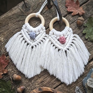 May include: Two macrame wall hangings with wooden rings and white fringe. Each features a central knot in either blue or pink. The hangings are displayed on a rustic wooden surface, with leaves and acorns scattered around.