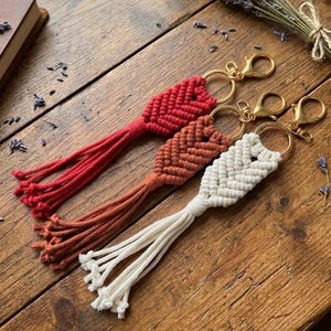 May include: Three macrame keychains in red, rust, and white, each with a gold-colored clasp and ring. The keychains are arranged on a wooden surface, with a book and dried lavender in the background. The keychains have a woven design and long tassels.