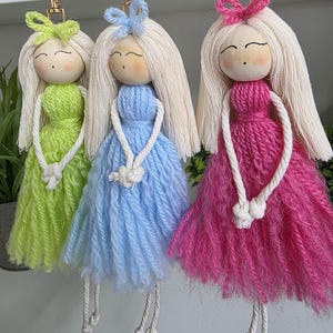May include: Three handmade doll keyrings with wooden heads, yarn hair, and colourful yarn dresses. The dolls have light green, blue, and pink dresses, with matching bows. Each doll has a white rope necklace and legs, and a gold keyring.