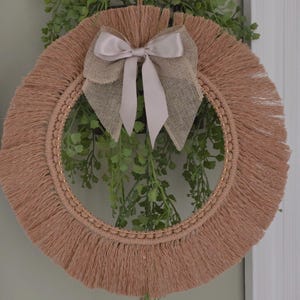 Puede incluir: Una corona de macramé redonda, beige, con borde de flecos y un lazo decorativo. El lazo está hecho de una combinación de arpillera y cinta de raso. Se ve vegetación verde detrás de la corona, añadiendo un toque natural.