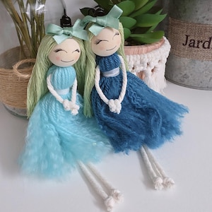 May include: Two handmade yarn dolls with smiling faces, green hair, and blue dresses. Each doll has a light green bow and white rope legs. The dolls are displayed on a white surface.