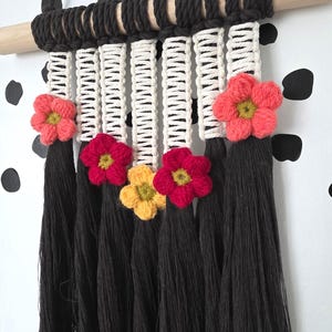 Puede incluir: Un tapiz de macramé con una barra de madera envuelta en cordón marrón oscuro. El diseño incluye secciones tejidas de color crema, borlas negras y flores de hilo hechas a mano en tonos rojos, rosas y amarillos. Una pieza decorativa.