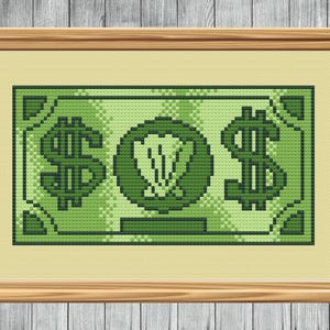 May include: A framed cross-stitch artwork depicting a green dollar bill. The design features large dollar signs on either side of a central emblem. The artwork is framed in a light brown wooden frame.