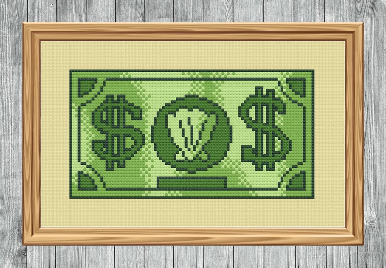 May include: A framed cross-stitch artwork depicting a green dollar bill. The design features large dollar signs on either side of a central emblem. The artwork is framed in a light brown wooden frame.