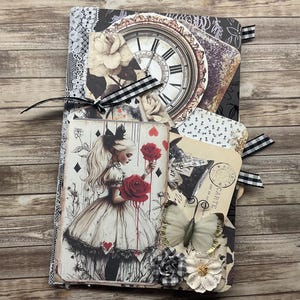 May include: A handmade journal with an Alice in Wonderland theme. The cover features a collage of images, including a clock, roses, and a playing card with a drawing of Alice holding roses. The journal is embellished with ribbons, lace, and paper flowers.