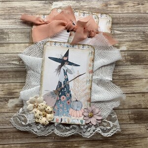 May include: A decorative tag featuring a printed illustration of a witch in a floral dress holding a broomstick next to pumpkins. The tag is layered with white cheesecloth, a peach-colored fabric ribbon bow, and small fabric flowers at the base.