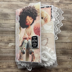 May include: A handmade journal with a decorative cover featuring a whimsical illustration of a girl with curly hair. The journal includes a pocket with a dog image, a bird, and a butterfly. The text "Find the Beauty in Every Day" is on the cover.