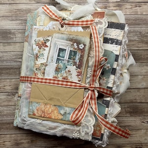 May include: A handmade junk journal featuring layered paper ephemera, floral prints, and a brown paper envelope. The book is bound with white lace trim and tied with an orange and white gingham ribbon, showcasing a mix of vintage-style textures.