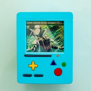 Può includere: Una console per videogiochi in plastica blu e nera con uno schermo che mostra una carta di Magic: The Gathering con Savra, Regina dei Golgari. La carta ha uno schema di colori verde, nero e bianco e raffigura una donna con lunghi capelli verdi che tiene uno scettro.