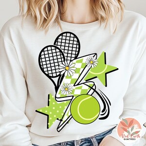 May include: White sweatshirt with a graphic design of two tennis rackets, a lightning bolt, a star, and a tennis ball. The design is in green, black, and white.