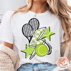 May include: White t-shirt with a graphic design of tennis rackets, a lightning bolt, stars, and a tennis ball. The design is in black, white, and green.