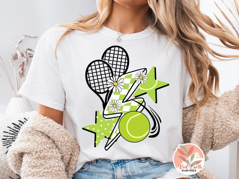 May include: White t-shirt with a graphic design of tennis rackets, a lightning bolt, stars, and a tennis ball. The design is in black, white, and green.
