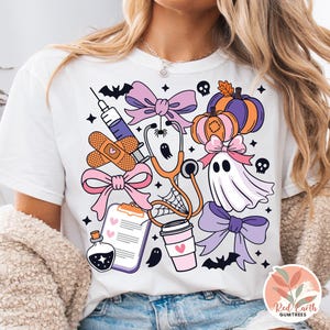 May include: White t-shirt featuring a colorful Halloween-themed design. The graphic includes a stethoscope, syringe, pumpkins, bows, a ghost, and bats. The design has a medical theme with a spooky twist.