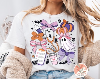 May include: White t-shirt featuring a colorful Halloween-themed design. The graphic includes a stethoscope, syringe, pumpkins, bows, a ghost, and bats. The design has a medical theme with a spooky twist.
