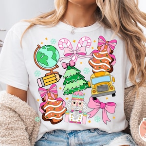 May include: White t-shirt featuring colorful Christmas-themed illustrations. Designs include a globe, pencil, Christmas tree, gingerbread cookies, school bus, candy canes, nutcracker, and bows. The shirt has a round neck and short sleeves.