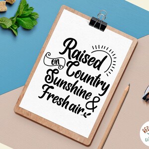 May include: A white clipboard with a black wood frame, featuring a handwritten quote that reads "Raised on Country Sunshine & Fresh Air." The quote is in a decorative script font. The clipboard is on a light brown surface.