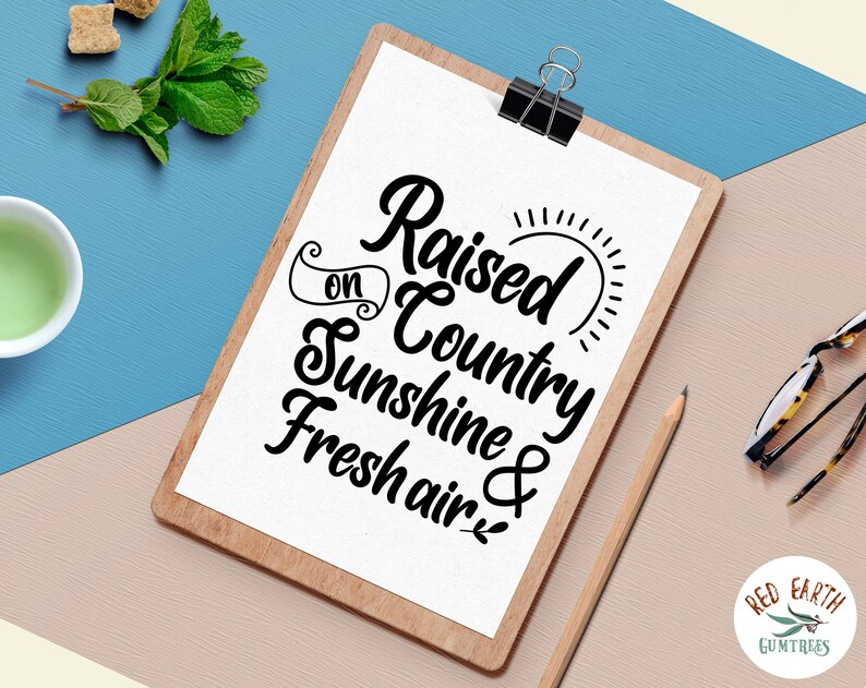 May include: A white clipboard with a black wood frame, featuring a handwritten quote that reads "Raised on Country Sunshine & Fresh Air." The quote is in a decorative script font. The clipboard is on a light brown surface.