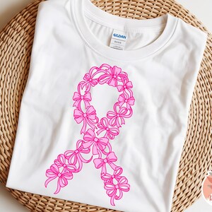 May include: White t-shirt featuring a pink ribbon design made of bows. The shirt is displayed on a woven, light brown surface. The design is a symbol of breast cancer awareness.