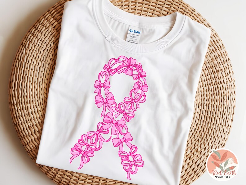 May include: White t-shirt featuring a pink ribbon design made of bows. The shirt is displayed on a woven, light brown surface. The design is a symbol of breast cancer awareness.