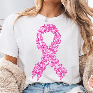 May include: White t-shirt featuring a pink ribbon design made of bows. The ribbon is a symbol of breast cancer awareness. The shirt is a casual style, suitable for everyday wear. The design is centered on the front of the shirt.