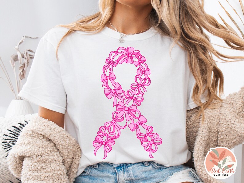 May include: White t-shirt featuring a pink ribbon design made of bows. The ribbon is a symbol of breast cancer awareness. The shirt is a casual style, suitable for everyday wear. The design is centered on the front of the shirt.
