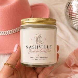 May include: A glass jar candle with a gold lid, labelled "Nashville Bachelorette." The label features a cowboy hat and boots illustration. The text reads "Smells like Broadway and bad decisions." A pink cowboy hat is in the background.