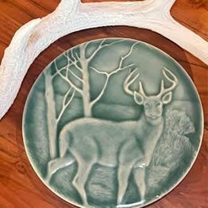 May include: A teal ceramic plate featuring a detailed relief of a deer in a forest scene. The deer has large antlers and is surrounded by trees. The plate is round and has a glossy finish. A white antler is in the background.