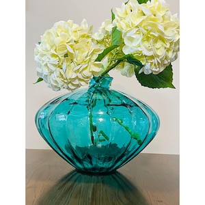 May include: A teal glass vase with a unique, ribbed design, holding a bouquet of creamy white hydrangeas. The vase has a rounded shape and sits on a dark wooden surface, creating a striking contrast.