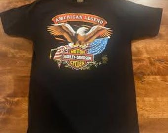May include: Black t-shirt featuring a graphic of a bald eagle with outstretched wings, the American flag, and the text "AMERICAN LEGEND" and "HARLEY-DAVIDSON CYCLES". The design is in shades of red, white, blue, and gold.