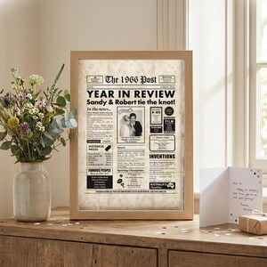 May include: A framed print of a newspaper page titled "The 1966 Post" with the headline "YEAR IN REVIEW." The article highlights Sandy & Robert's wedding. A vase of flowers and a greeting card are on a wooden surface.