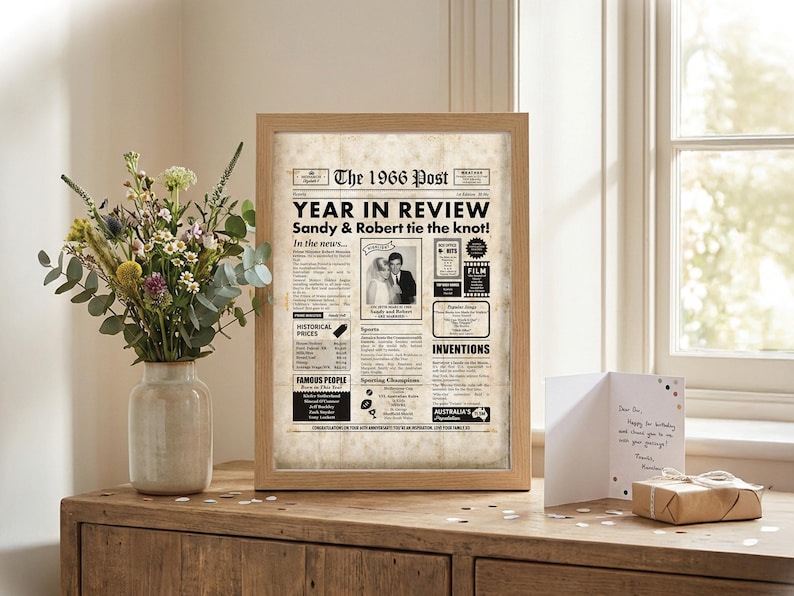 May include: A framed print of a newspaper page titled "The 1966 Post" with the headline "YEAR IN REVIEW." The article highlights Sandy & Robert's wedding. A vase of flowers and a greeting card are on a wooden surface.