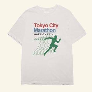 May include: White t-shirt with the text "Tokyo City Marathon" in red and blue, and Japanese text. A green runner silhouette with motion lines suggests speed. The design has a vintage aesthetic, suitable for sports enthusiasts or fans of Tokyo.