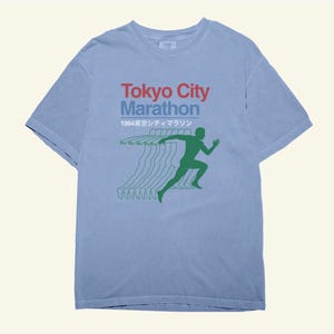 May include: A pale blue t-shirt with the words "Tokyo City Marathon" in red and blue, and a green running figure graphic. The shirt also features the year "1994" and Japanese text. The design is printed on a short-sleeved tee.