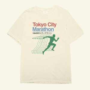 May include: A cream-coloured t-shirt with the words "Tokyo City Marathon" in red and blue, and Japanese text below. A green silhouette of a runner is shown, with green lines trailing behind. The design has a vintage aesthetic.