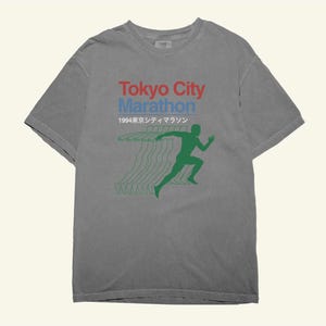 May include: Grey t-shirt featuring the text "Tokyo City Marathon" in red and blue. Below is "1994" in Japanese characters. A green runner silhouette is printed on the front, with green lines trailing behind.