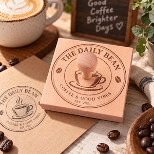 Può includere: Un timbro quadrato in legno con impugnatura caratterizzato da un design circolare inciso che riporta la scritta THE DAILY BEAN, COFFEE & GOOD VIBES, EST. 2021, con un'illustrazione centrale di una tazzina e icone di chicchi di caffè.