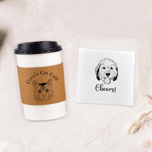 May include: A white coffee cup with a brown sleeve that says "Coco's Cat Cafe" and a black and white illustration of a cat. A white napkin with a black and white illustration of a dog and the text "Cheers!"