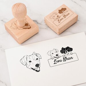 May include: A wooden rubber stamp with two black and white dogs peeking over a banner that reads "Liou Nhan". The stamp is on a white surface.