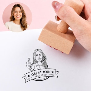 May include: A wooden rubber stamp creating a black ink impression on white paper. The design features a line art illustration of a person giving a thumbs up, with a banner below containing the text "GREAT JOB! KEEP IT UP!" and two stars.