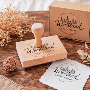 Puede incluir: Un sello de goma rectangular de madera con mango, que presenta un logotipo circular con pinos, una pequeña cabaña y el texto Woodland CRAFT STUDIO. El sello va acompañado de una caja de cartón a juego y un cuadrado de papel estampado.
